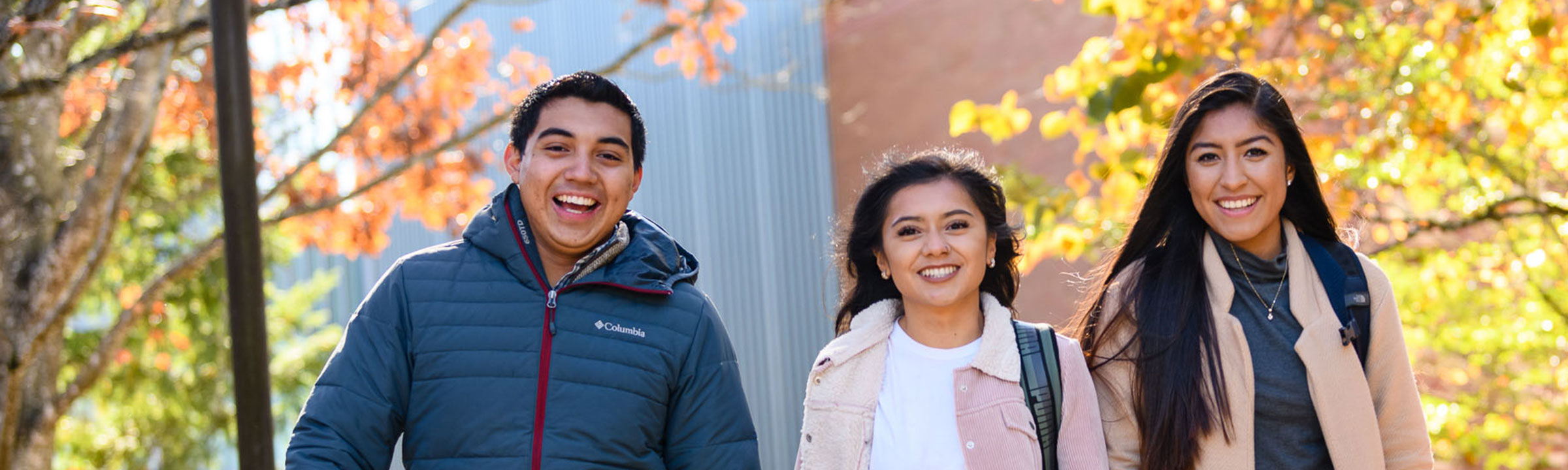 Three students smiling and walking on campus in front of campus building and fall leaves