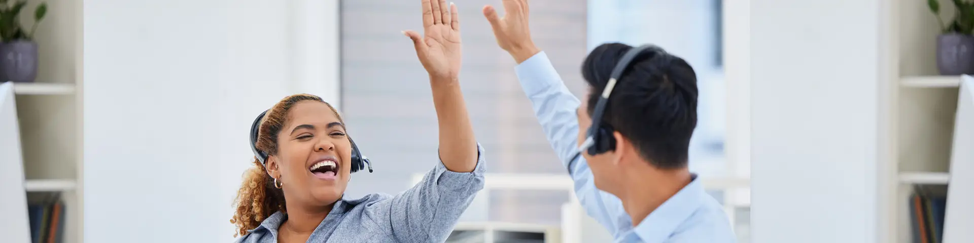 Two office workers in a corporate setting wearing headsets smile and high five