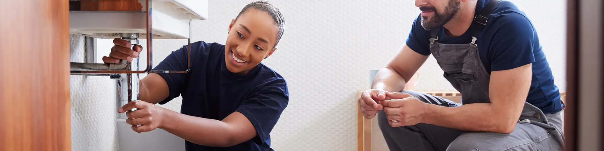 Plumbing apprentice works on bathroom sink pipe with journeyman observing