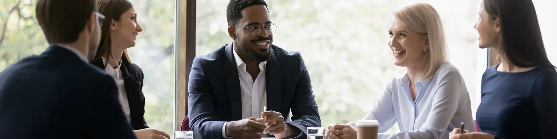 A group of five business professionals in formal attire speaking together at a large table during a business meeting.