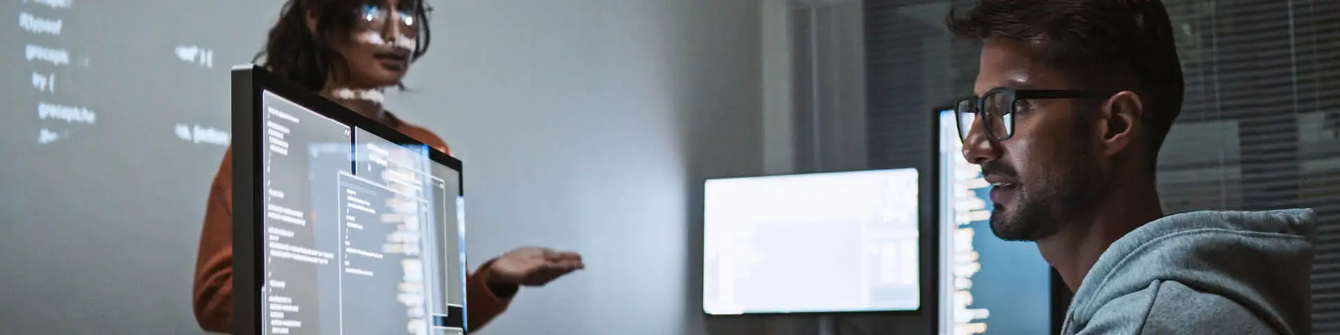 Two people: one is looking at a monitor projecting lines of code on a wall; one standing in front.
