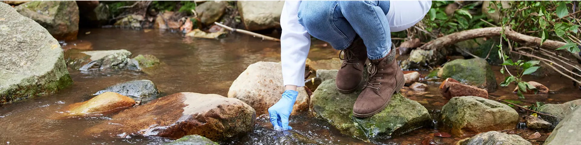 Geology student with a white lab coat bending over to gather a water sample from a river.