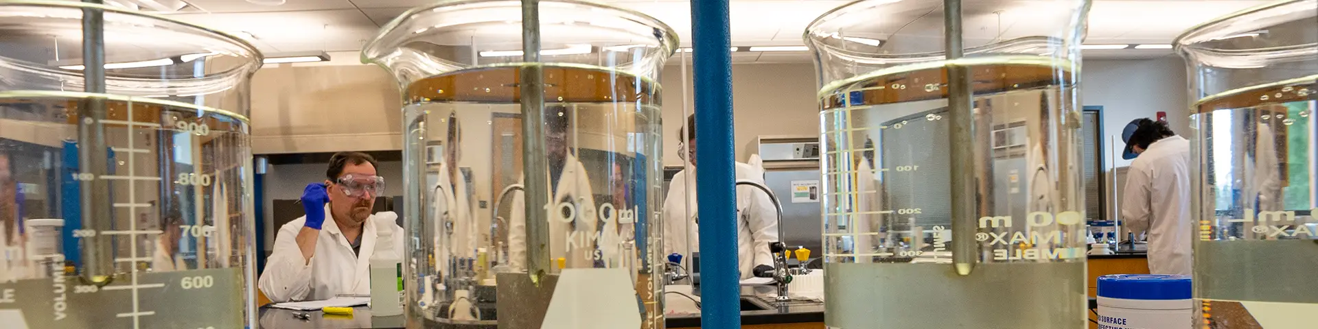 Chemistry lab: Students in white lab coats and protective eyewear can be seen through beakers of clear liquid in the foreground