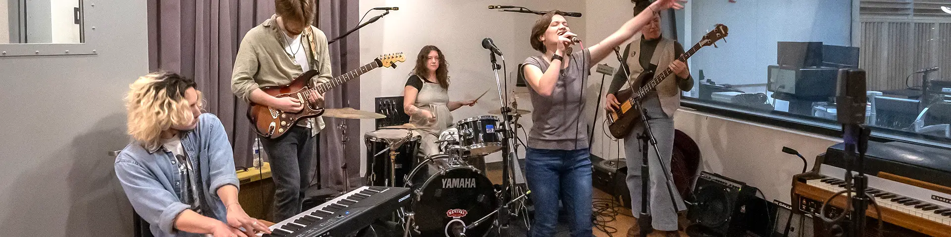 A student ensemble, including students on keyboard, electric guitar, drum set, bass and vocals, practices in a CCC music studio.