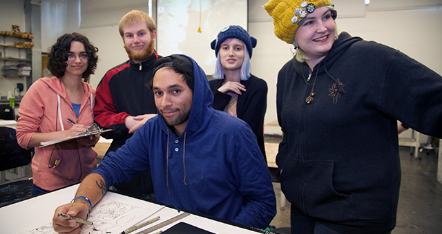 Student in blue hoodie draws at a table in a classroom surrounded by four other students, including a student with blue hair and another wearing a hat decorated with pins