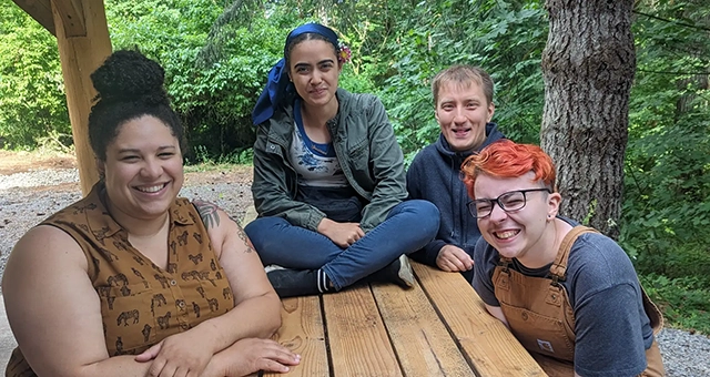 Three CCC students and a staff member smile around a picnic table at an outdoor gathering, with one student sitting cross-legged on the table