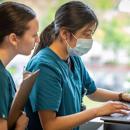 Two healthcare providers in scrubs, one wearing a surgical mask, work on a laptop