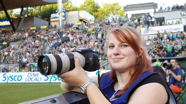 Student photo journalist poses with a camera in the Timbers press box at Providence Park